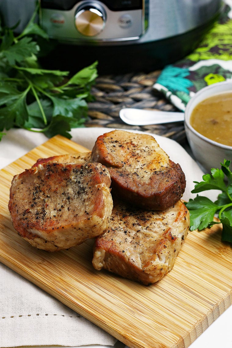 A wooden cutting board of pork chops, a bowl of gravy, and an Instant Pot in the background.