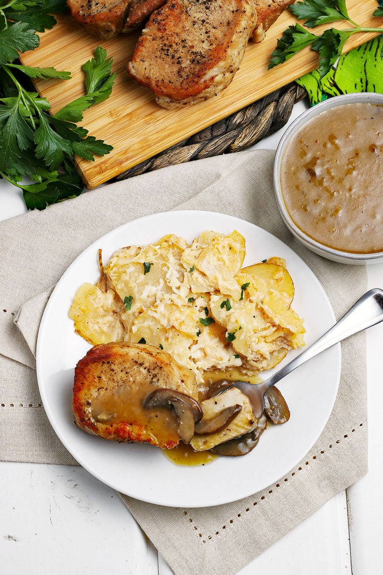 Overhead shot of pork chops and scallop potatoes on a white plate with fork on a linen napkin with a bowl of gravy with pork chops on a wooden cutting board in the background.
