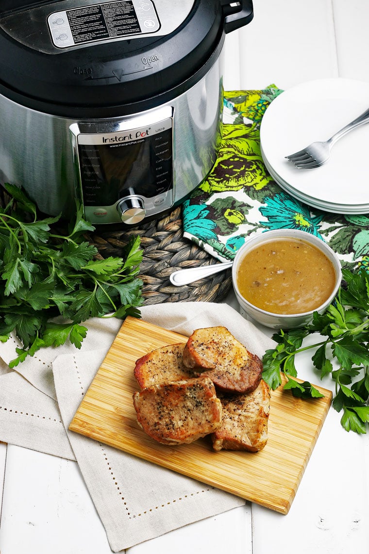 Pork chops on a wooden cutting board, a bowl of mushroom gravy, an Instant Pot on a woven mat, and white plates in the background.