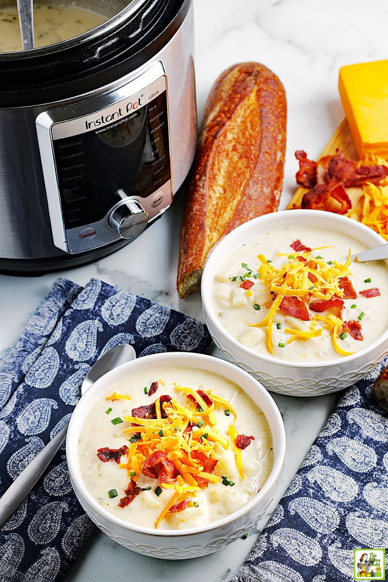 Overhead view of two bowls of loaded potato soup , blue napkins, a spoon, an Instant Pot, a small loaf of bread, and cheese.