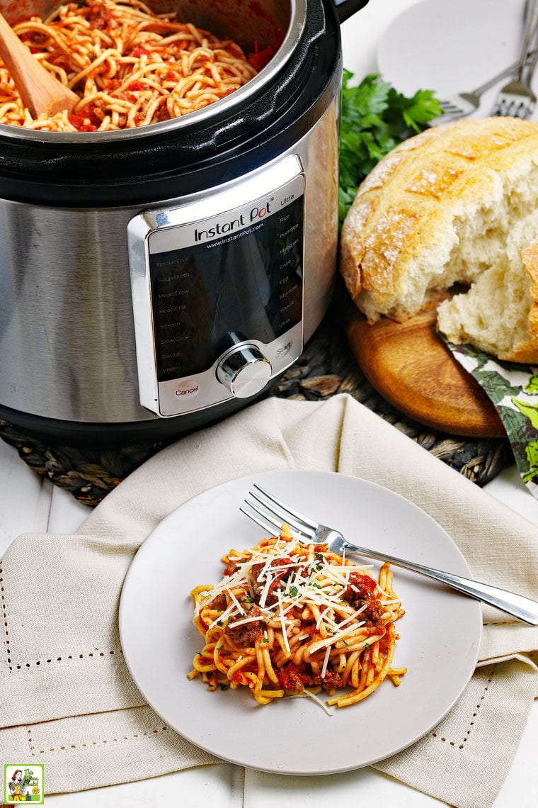 A white plate of spaghetti with meatsauce with a fork on a gray napkin with an Instant Pot of spaghetti with a wooden spoon, a loaf of crusty bread and a stack of plates in the background.
