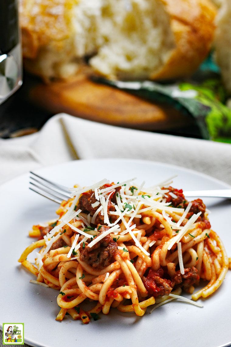 Closeup of a plate of spaghetti and meatsauce with a fork on a linen napkin with bread and a pressure cooker in the background.