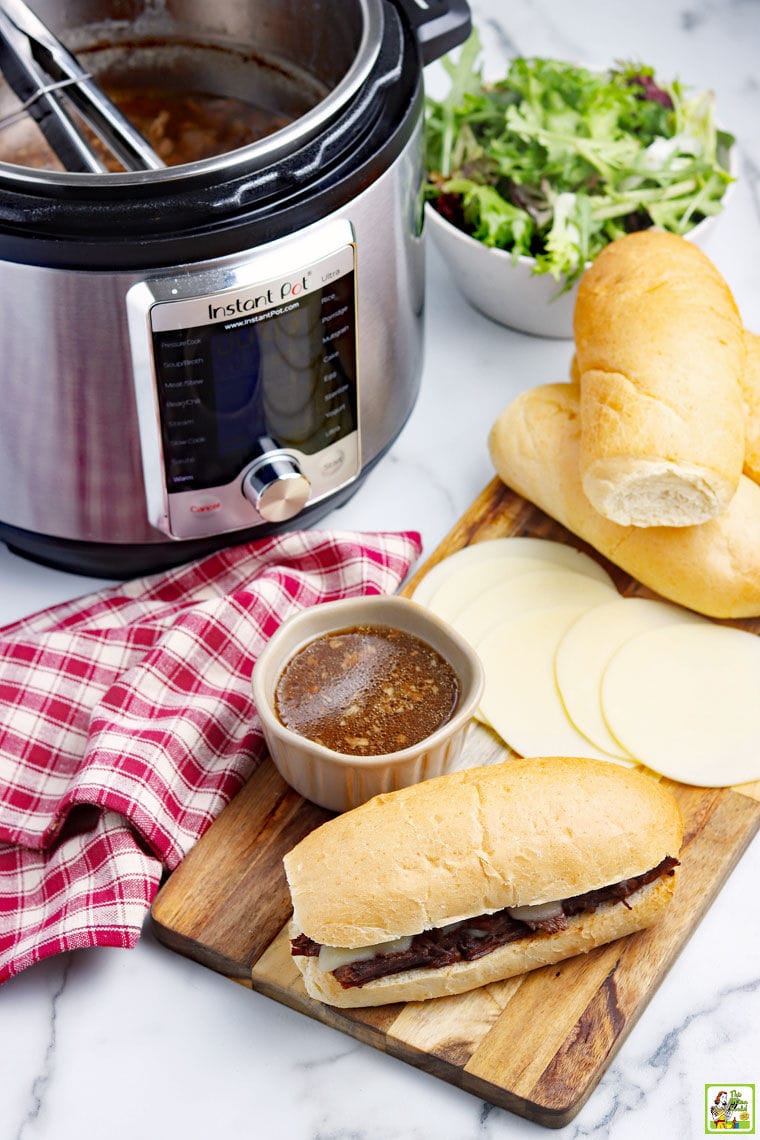 Overhead shot of a Instant Pot French Dip sandwich with bowl of au jus, red and white napkin, slices of cheese and bread rolls, an Instant Pot of French Dip, and a bowl of salad.