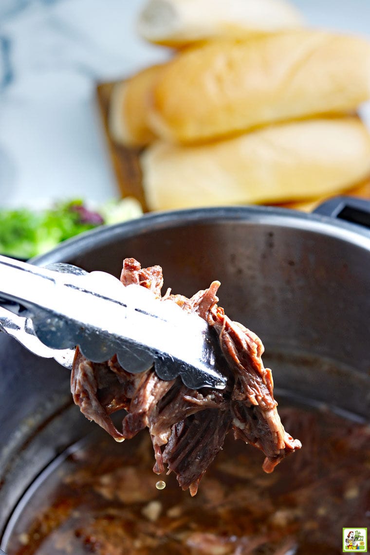 Piece of French DIp meat in tongs over a Instant Pot with sandwich rolls in the background.