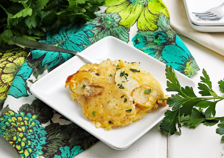 A white plate of Super Cheesy Crockpot Scalloped Potatoes with a fork on a floral napkin with fresh parsley.