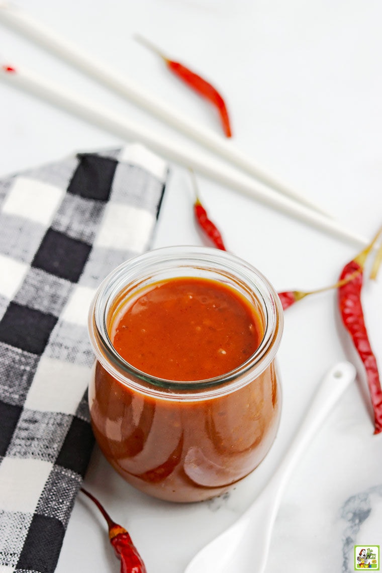 Overhead shot of glass jar of hoisin sauce with black and white napkin, red peppers, white spoon, and chopsticks.