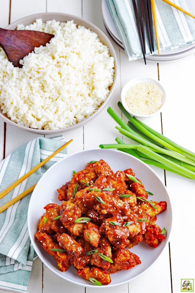 Overhead shot of a bowl of General Tso Chicken, a bowl of rice, green onions, chopsticks, napkins, and plates.