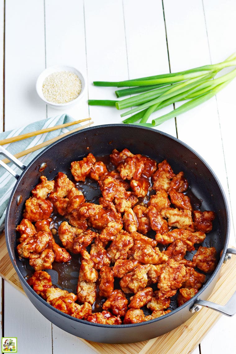 A frying skillet of cooked General Tso's chicken with chopsticks, green onions, and a small bowl of sesame seeds.