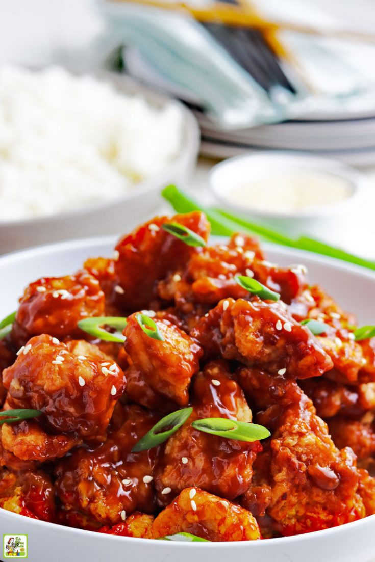 Closeup of a bowl of General Tso's chicken stir fry with sesame seeds and sliced green onions with a bowl of steamed rice in the background.