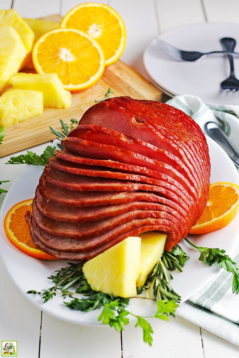 A glazed ham on a white ceramic platter with pineapple and orange slices.