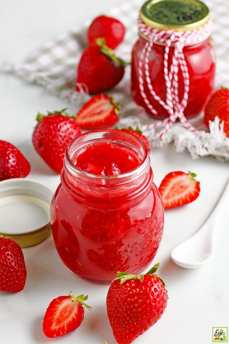 Glass jars of strawberry sauce with a serving spoon, gray dish towel, and sliced and whole strawberries.