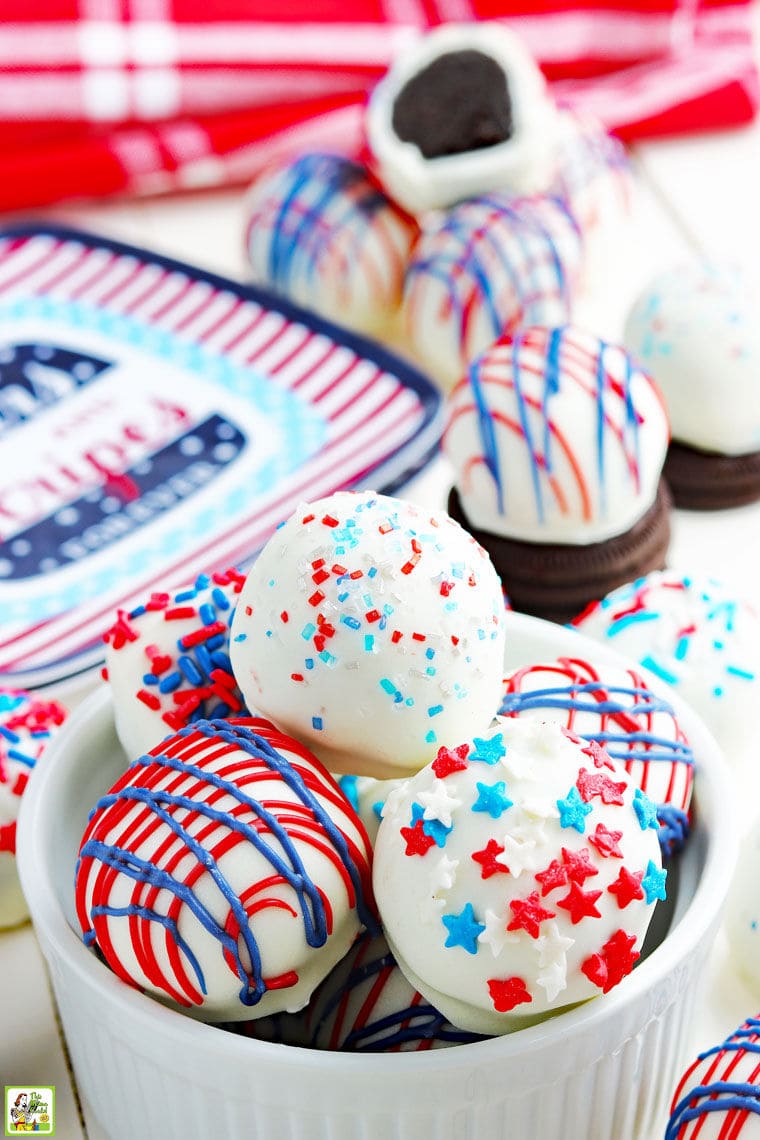 Red, white and blue Oreo cake balls in a bowl and with Oreos and holiday paperplates on a festive tabletop.