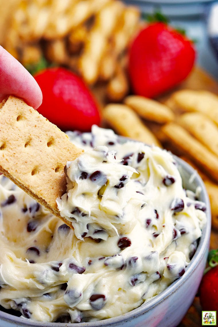 A hand dipping a piece of graham cracker into chocolate chip cookie dough in a small bowl with fruit, crackers, and cookies in the background.