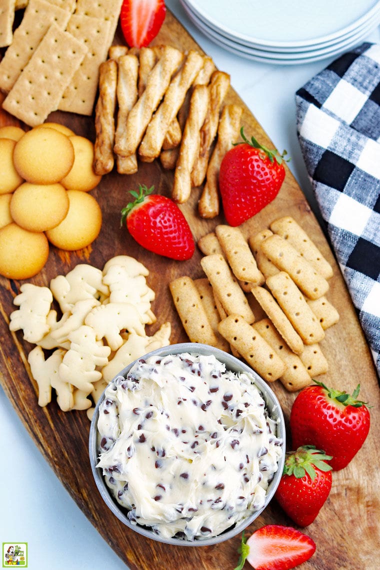 Overhead of dessert board of a bowl of chocolate chip cookie dough dip, graham crackers, fruit, nilla wafer cookies, and pretzels with a black and white napkin.