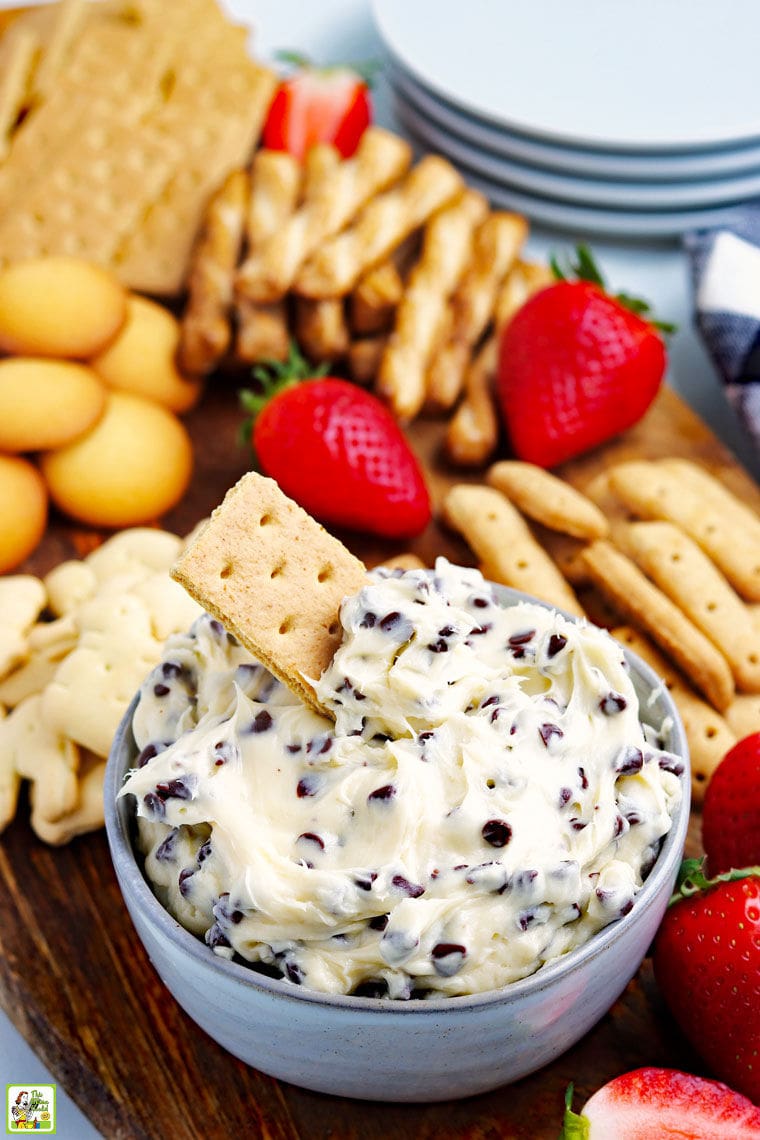 A bowl of cookie dough dip recipe with a graham cracker with more dippers like cookies, crackers, and fruit and plates in the background.