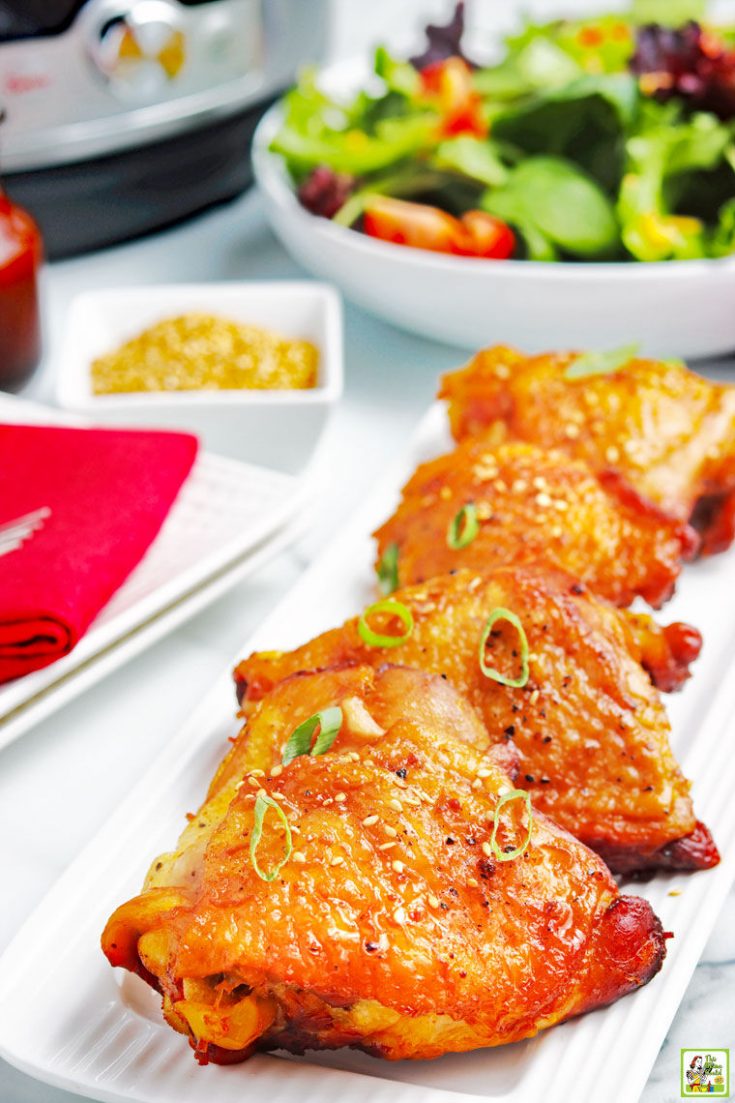 Four pieces of Instant Pot Chicken Thigh with Honey Garlic Sauce on a white platter with a bowl of salad, bowl of Dijon mustard, red napkin, plates, and pressure cooker in the background.