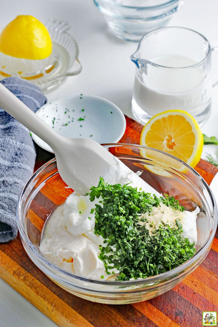 Making Low-Carb Keto Ranch Dressing in a bowl with fresh herbs, lemons, and small bowls on a wooden cutting board with a dish towel.