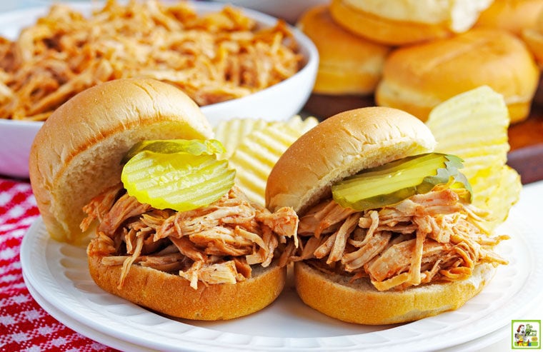 Two Instant Pot Pulled BBQ Chicken Sandwiches with pickles and potato chips on a white plate with bowl of shredded chicken and buns in the background.