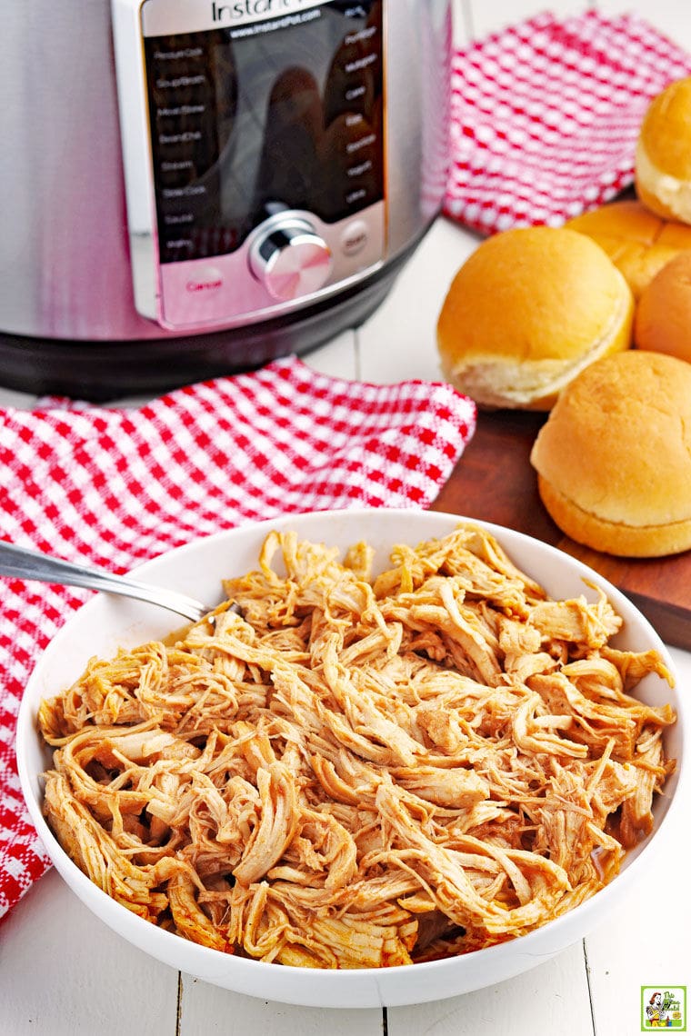 A bowl of shredded Instant Pot Pulled BBQ Chicken with fork, sandwich buns, red checkered napkins, and a pressure cooker.