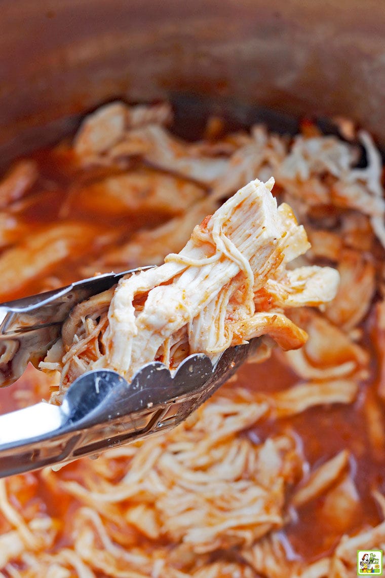 Closeup of shredded chicken in tongs above pressure cooker filled with Instant Pot Pulled BBQ chicken.