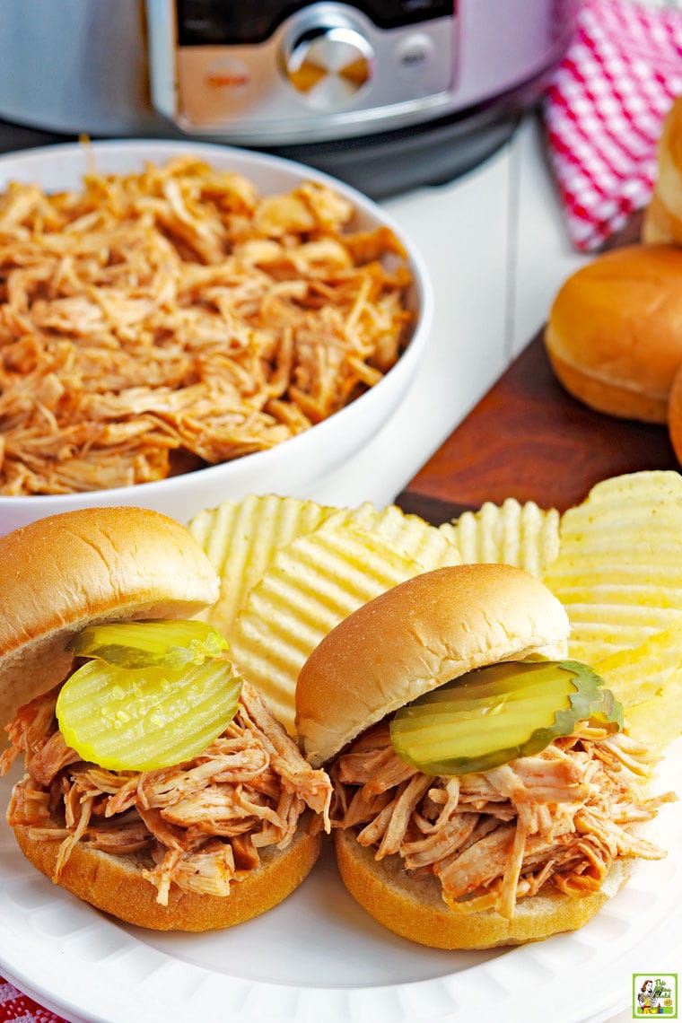 Two Instant Pot Pulled BBQ Chicken Sandwiches with potato chips on a white plate with a bowl of shredded BBQ chicken with a pressure cooker, sandwich buns, and a red checkered napkin.