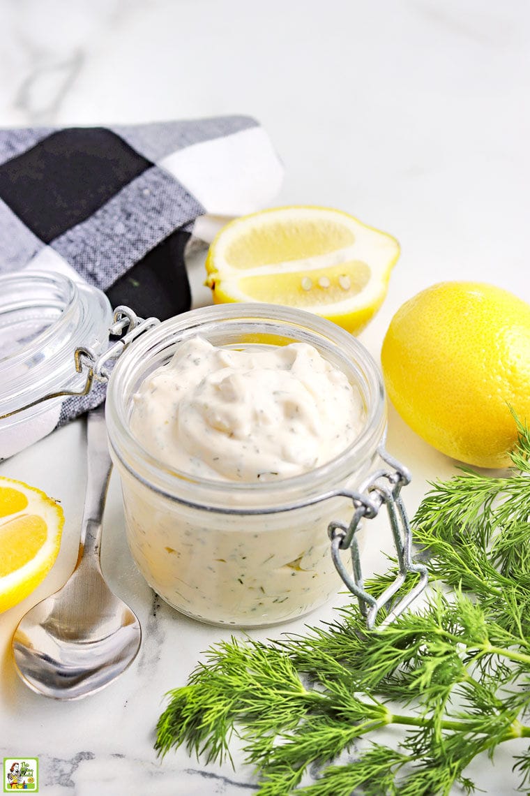 A glass jar filled with homemade tartar sauce, a bunch of fresh dill, lemons, a spoon, a whisk, and a black and white cloth napkin.