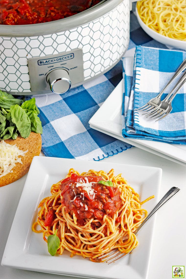 A white plate of crockpot spaghetti with meat sauce with blue and white napkins with plates and a slow cooker of sauce.