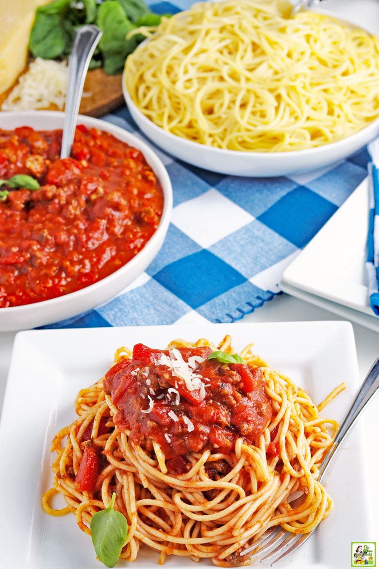 Overhead view of a plate of spaghetti sauce on pasta with bowls of crockpot spaghetti sauce and pasta with blue and white checked napkins.