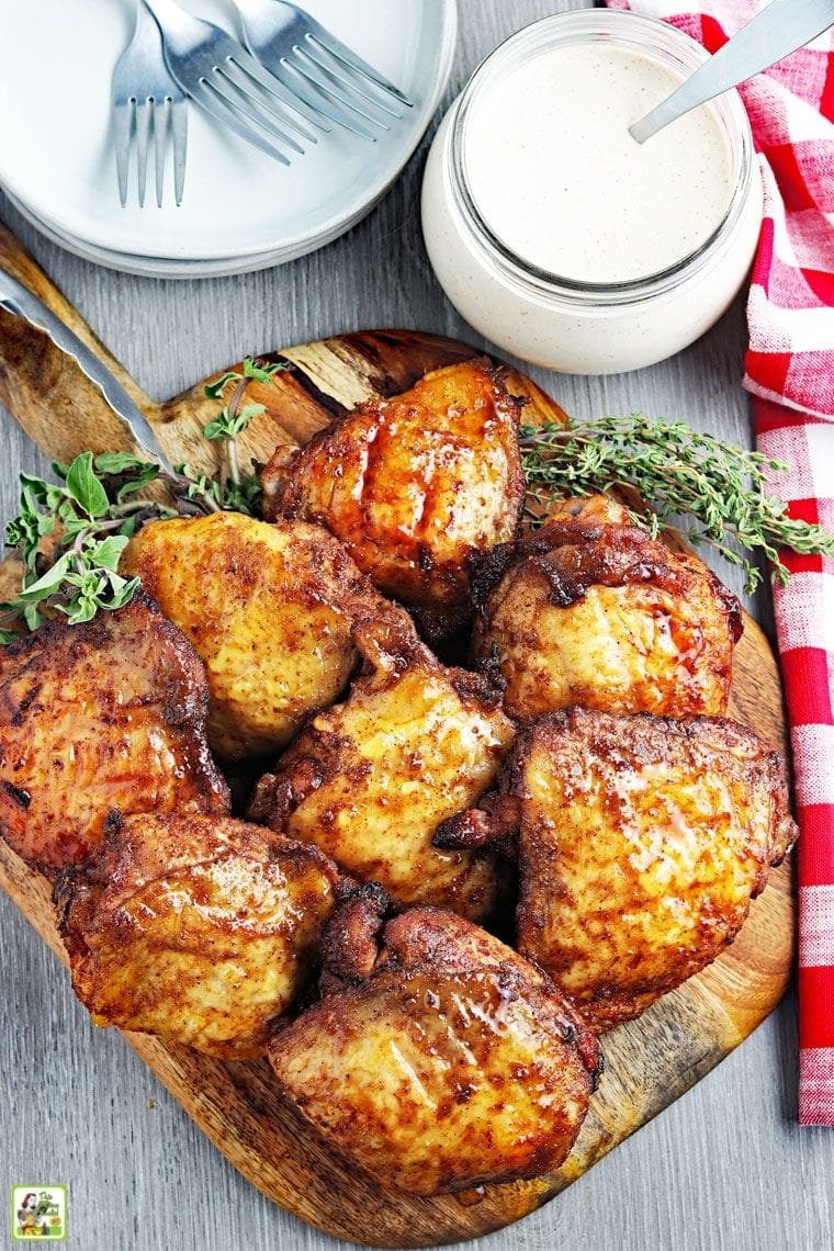 Overhead view of cooked smoked chicken thighs on a wooden cutting board with a jar of Alabama sauce, plates, utensils, and red and white cloth napkins.