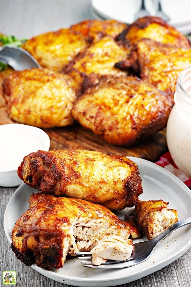 A plate of smoked chicken thighs with more chicken in the background and jars of white dipping BBQ sauce.