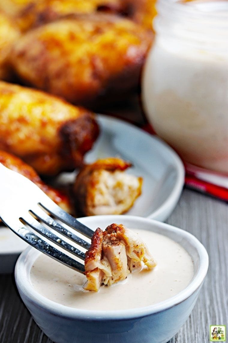 A piece of cooked chicken on a fork being dipped into Alabama barbecue sauce with pieces of cooked smoked chicken thighs in the background.