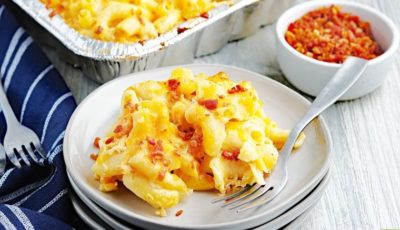 A serving of smoked mac and cheese on a white plate with a fork, blue napkin, and bowl of bacon bits.