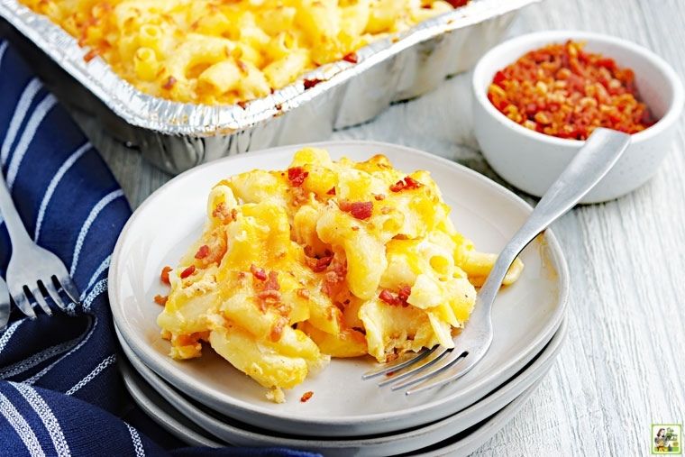 A serving of smoked mac and cheese on a white plate with a fork, blue napkin, and bowl of bacon bits.
