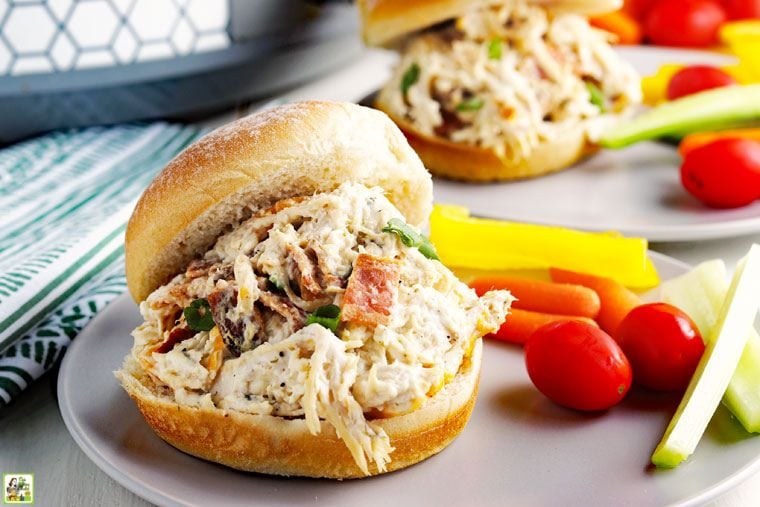 Crock pot crack chicken served on hamburger buns with cherry tomatoes and veggie sticks on a gray plate.