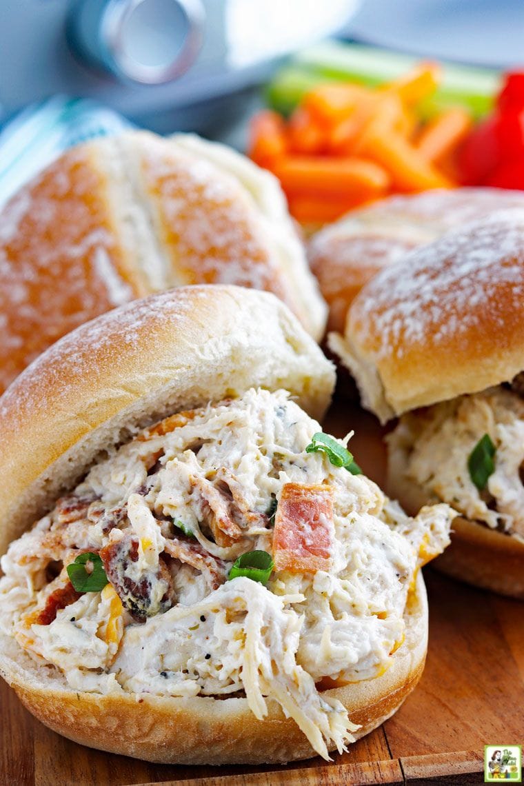 Three slow cooker crack chicken sandwiches served on hamburger buns on a wooden cutting board.