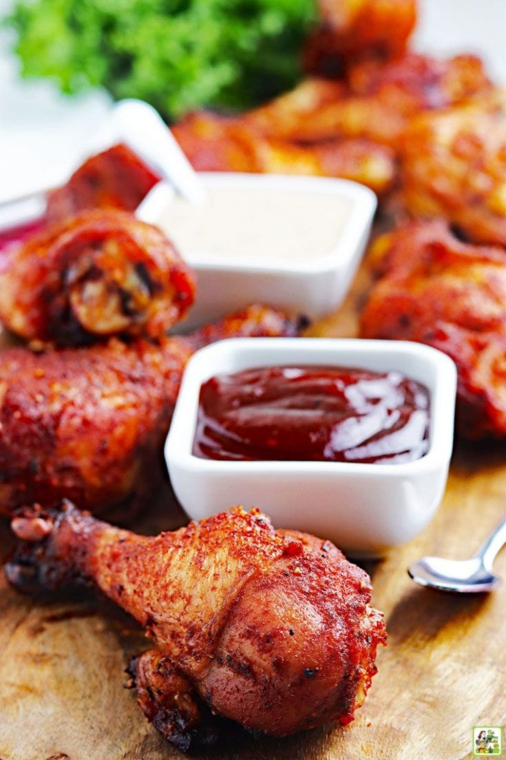 Smoked chicken legs with on a wooden board with small bowls of BBQ sauces.