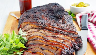 Sliced Traeger smoked brisket on a wooden cutting board with knife, BBQ sauce, garnish, and napkin.