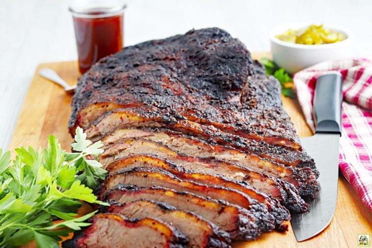 Sliced Traeger smoked brisket on a wooden cutting board with knife, BBQ sauce, garnish, and napkin.