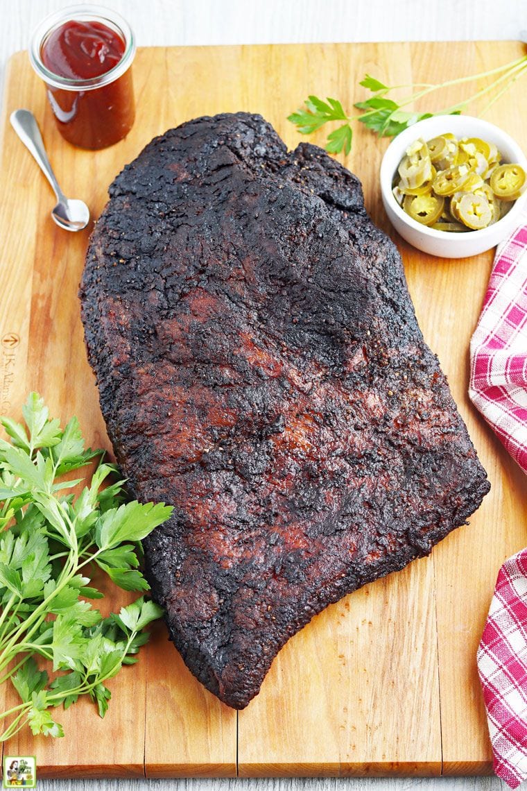 Smoked brisket on a wooden cutting board with jars of barbecue sauce and jalapenos.