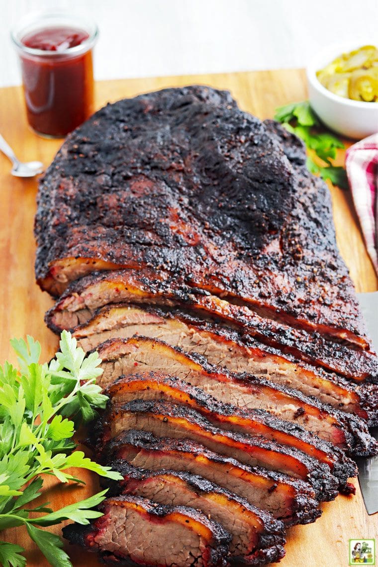 Sliced cooked beef brisket on a wooden cutting board with a jar of BBQ sauce.