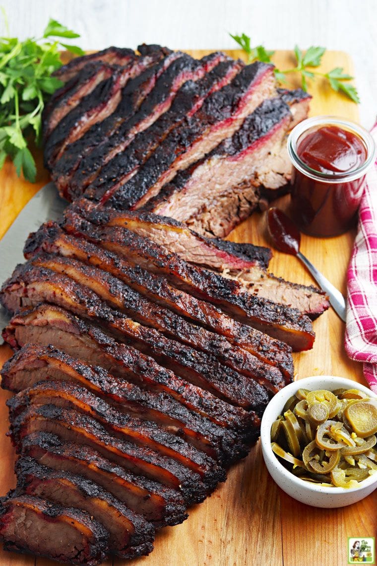 Serving sliced smoked brisket with barbecue sauce and jalapenos on a wooden cutting board with a red and white napkin. 