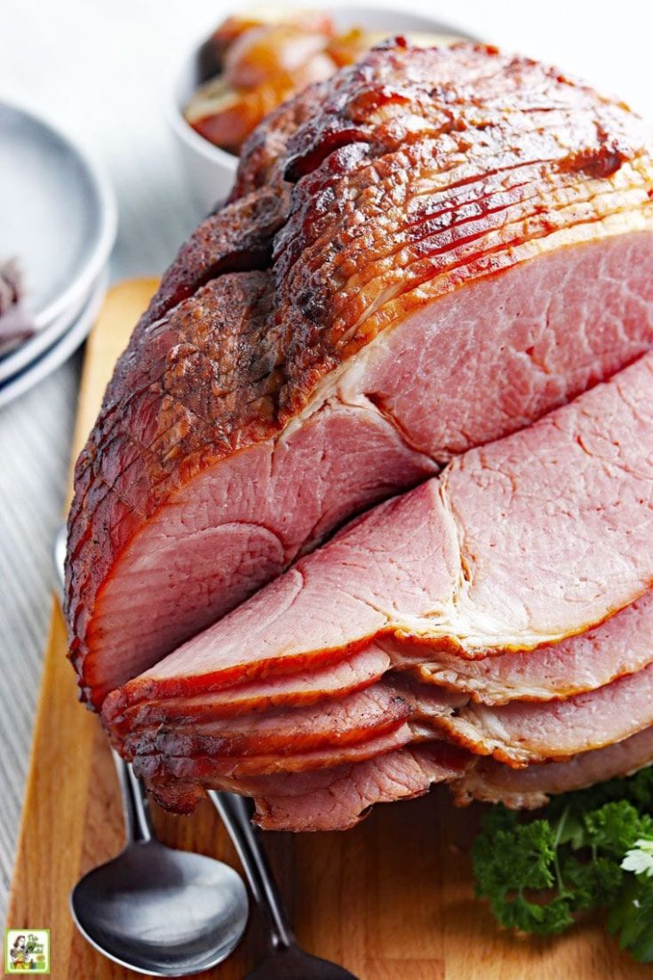 Sliced spiral cut ham with metal serving utensils on a wooden cutting board.