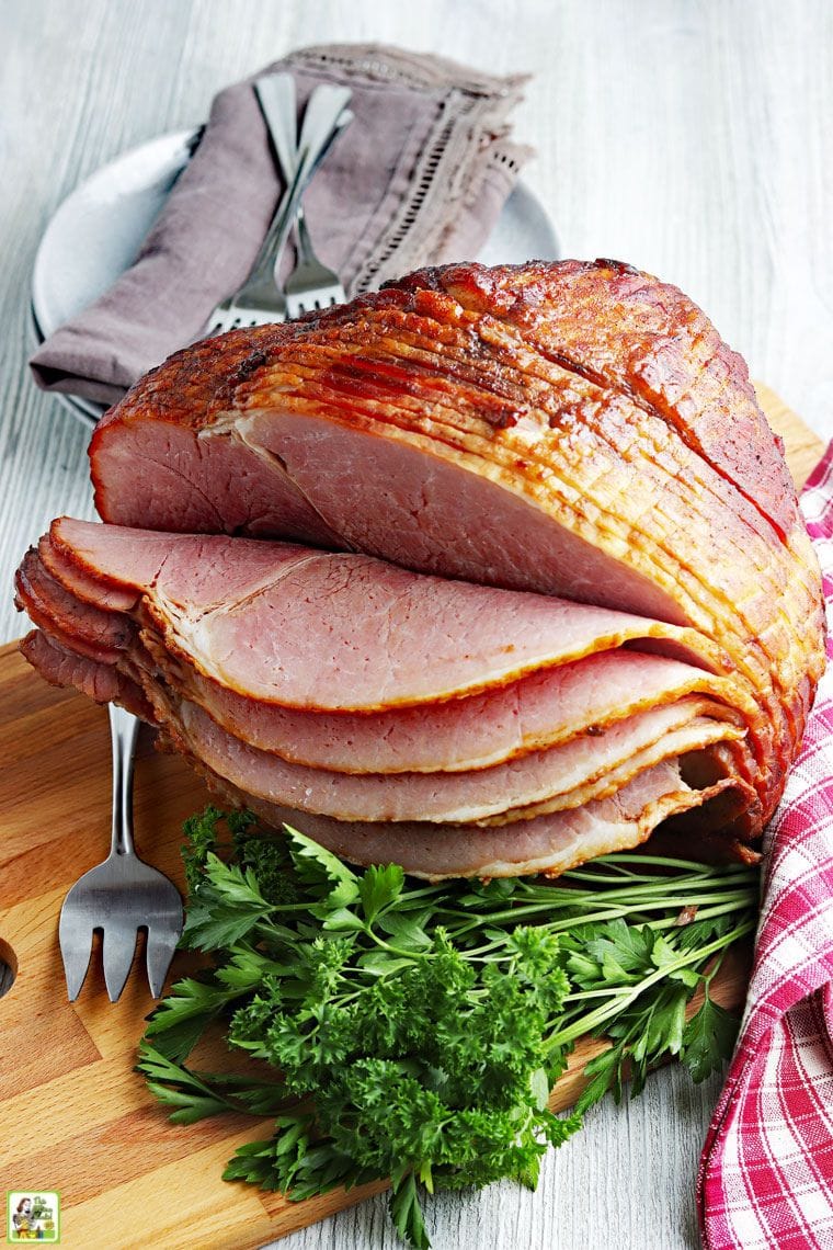 Glazed spiral cut bone in ham on a wooden cutting board with serving utensils, cloth napkin, and a bunch of parsley.