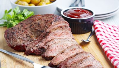 Sliced smoked tri-tip on a wooden cutting board with BBQ sauce, utensils, a bowl of roast potatoes, and a checkered napkin.