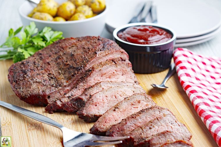 Sliced smoked tri-tip on a wooden cutting board with BBQ sauce, utensils, a bowl of roast potatoes, and a checkered napkin.