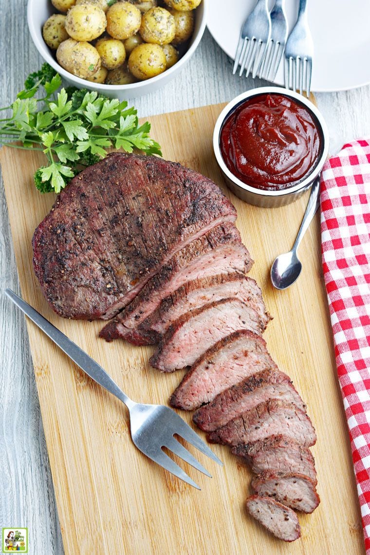 Overhead view of a sliced and cooked smoked tri-tip with serving fork and spoon, bowl of BBQ sauce, plates, bowl of potatoes, and cloth napkin.